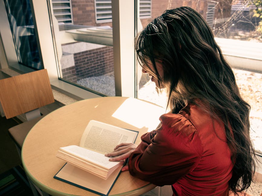 A young woman with long hair reads at a table by a sunlit window in a library.